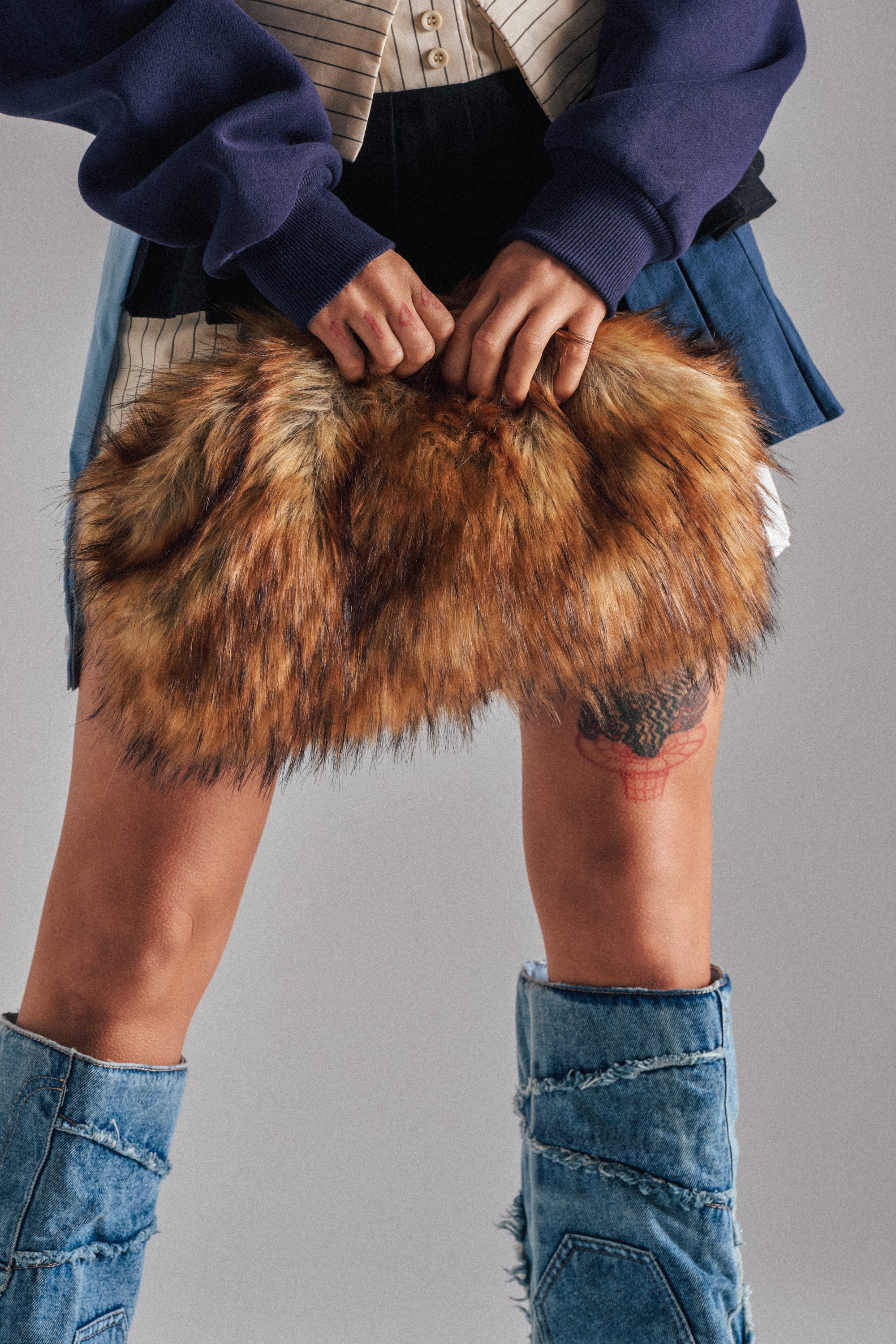 A person in a blue and white outfit holds the WILD CHILD FAUX FUR CLUTCH. Their leg tattoo and knee-high patchwork denim boots are visible, with the image cropped to show only their lower torso and legs. 1