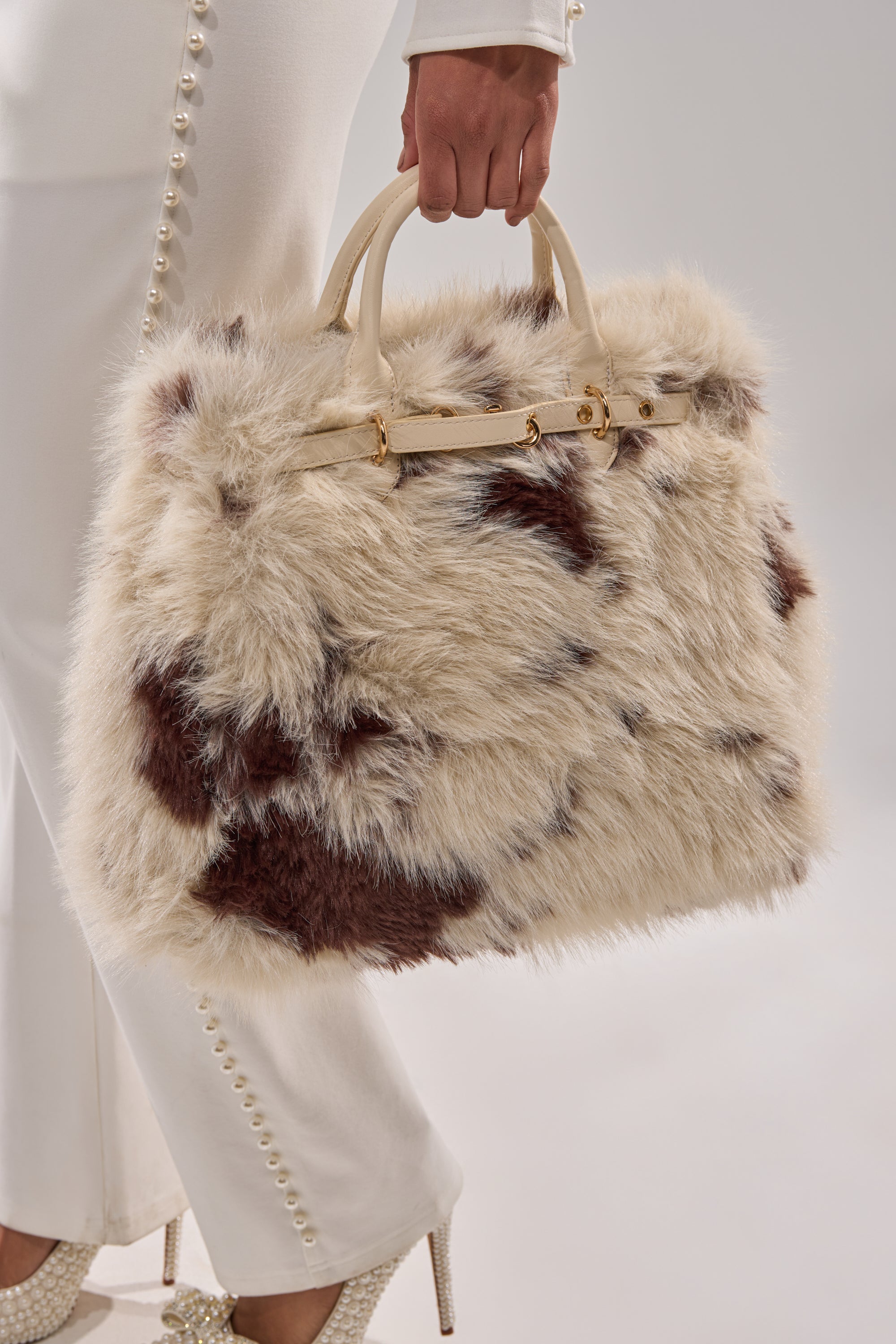 A person holds the MOO FAUX FUR MINI TOTE, a beige-and-brown cow print bag with cream handles, while wearing pearl-embellished white pants and sparkling heels against a light background.