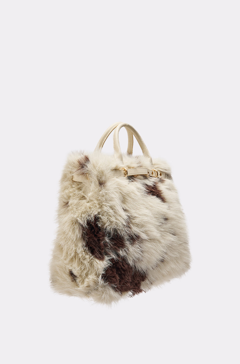 The MOO FAUX FUR MINI TOTE is a fluffy faux fur handbag with an off-white and brown cow print, two short handles, and a gold clasp, shown against a plain light background. 2