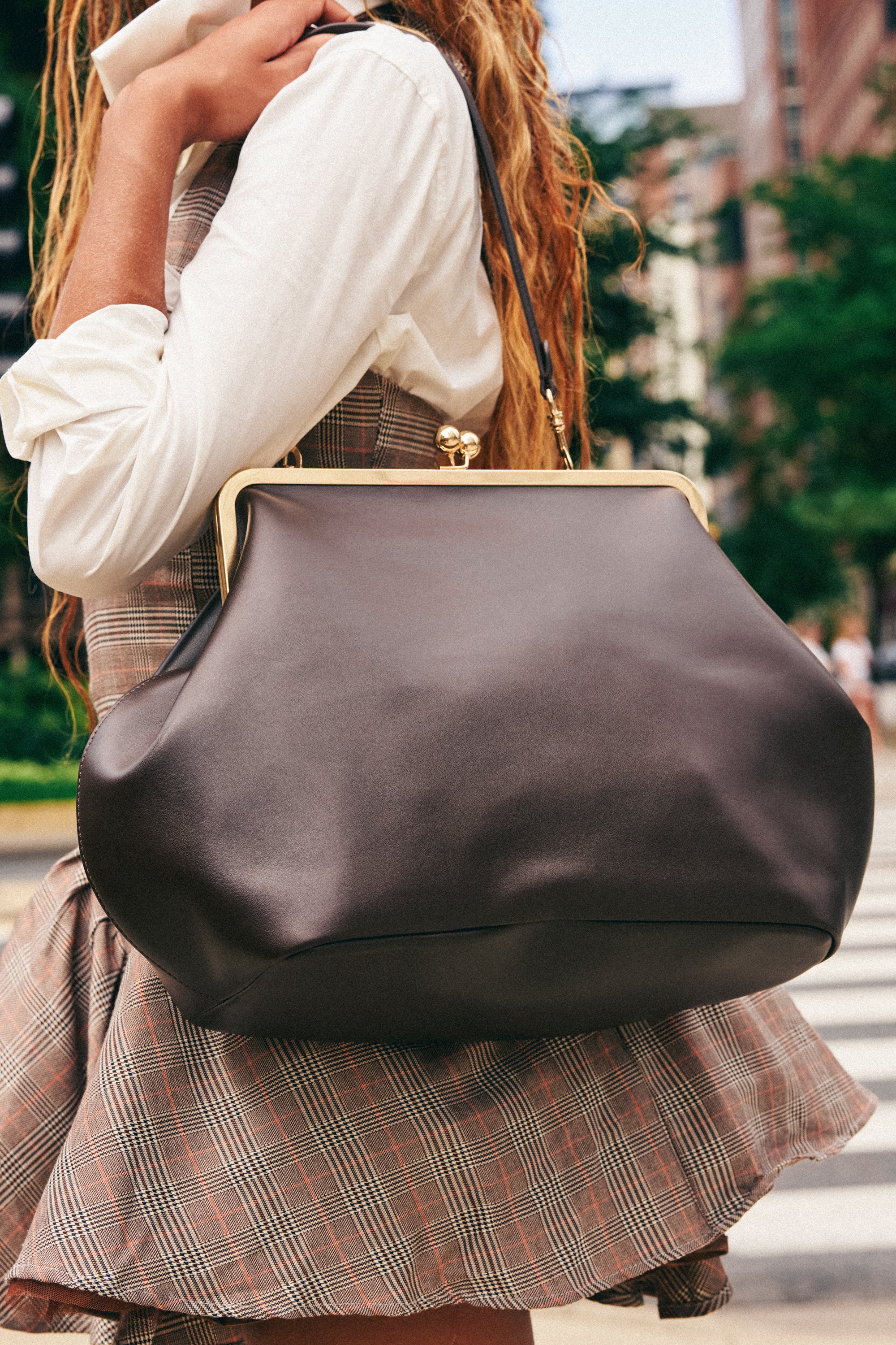 A woman in a plaid dress and white shirt holds the CLASSY GIRL BAG, an oversized clutch with gold hardware, while standing outdoors on a city street.