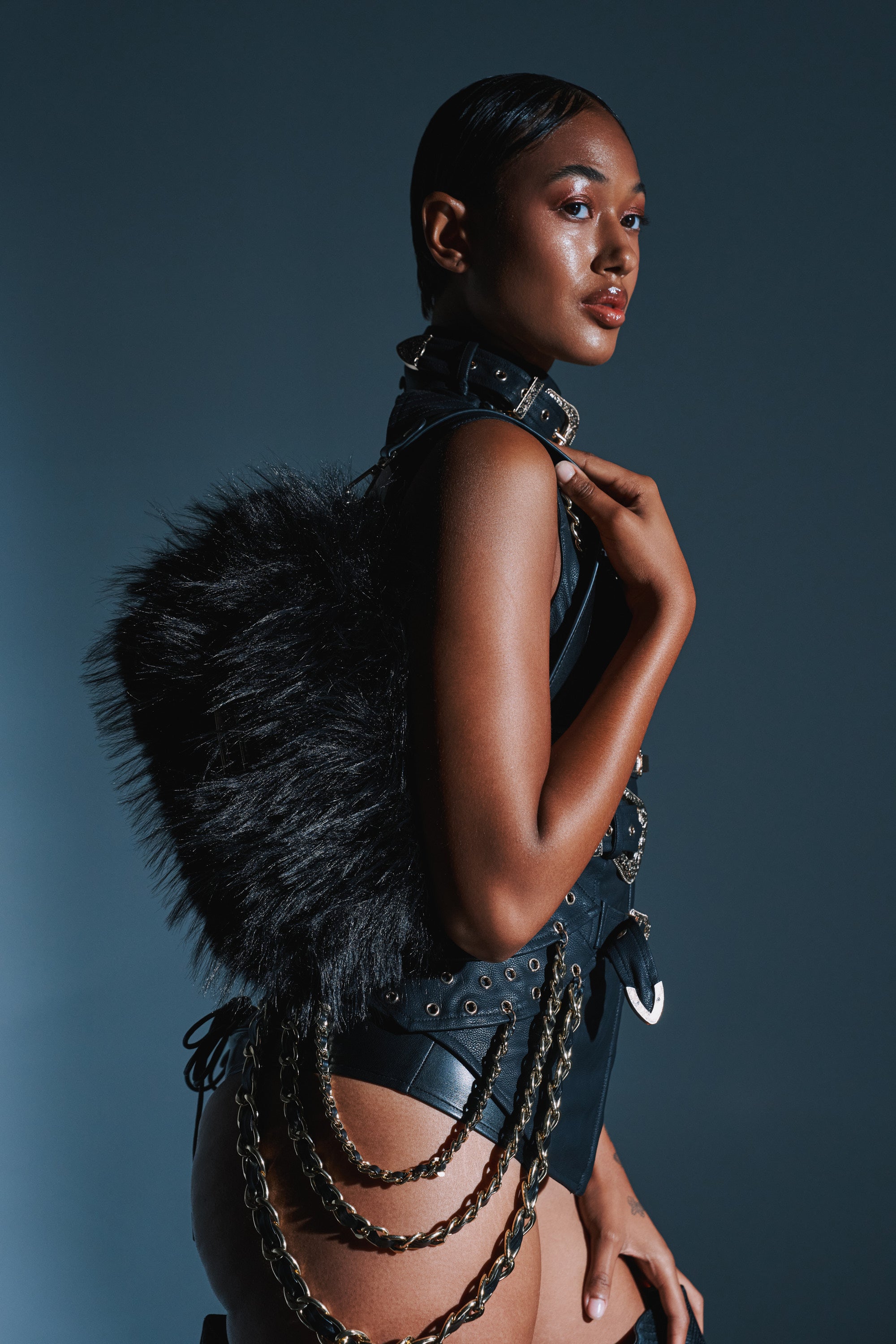 A person poses against a dark background, wearing a sleek black faux leather outfit with metal chains and carrying the REALLY LIKE THAT FAUX FUR PURSE IN BLACK on their back, confidently looking toward the camera.