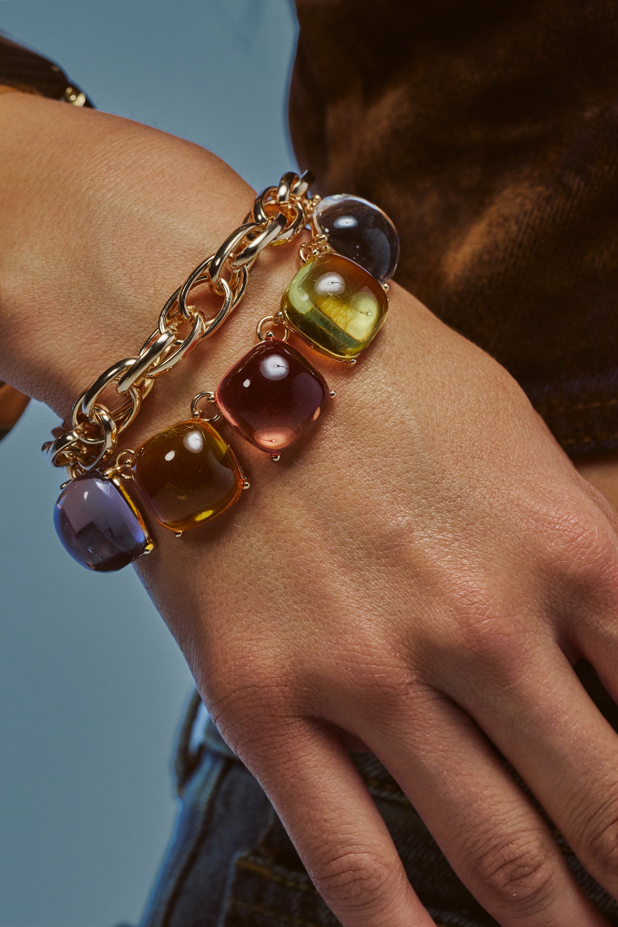 A hand wearing the MODERN MUSE STATEMENT BRACELET, featuring bold gold chain links and oversized pastel charms in purple, yellow, and amber. Styled with a brown top and jeans against a neutral blue background—perfect for showcasing vibrant summer accessories.