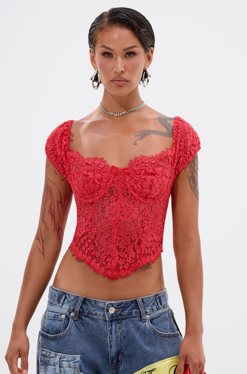 Person with short dark hair wears the GIRL ON FIRE LACE CORSET, blue jeans, hoop earrings, and a chain necklace. Tattoos are visible on their arms and chest as they stand against a plain white background.