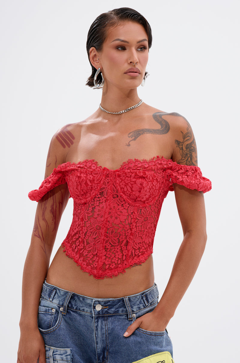 A person with short, slicked-back hair wears the GIRL ON FIRE LACE CORSET styled off-the-shoulder with blue jeans. They have tattoos on their arms and shoulders, hoop earrings, and a chain necklace, standing against a plain background.