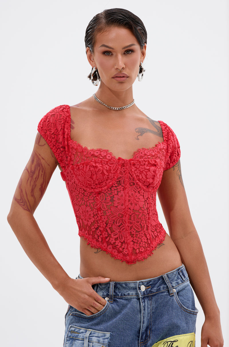 A person with slicked-back hair wears the GIRL ON FIRE LACE CORSET and blue patchwork jeans, confidently posing with one hand in their pocket against a white background. Tattoos on their arm and a chain necklace are visible.