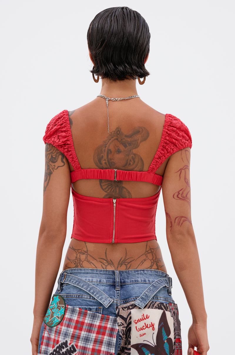A person with short dark hair, seen from the back, wears the GIRL ON FIRE LACE CORSET in red with ruched sleeves and blue patchwork jeans. Multiple tattoos are visible on their back and arms.