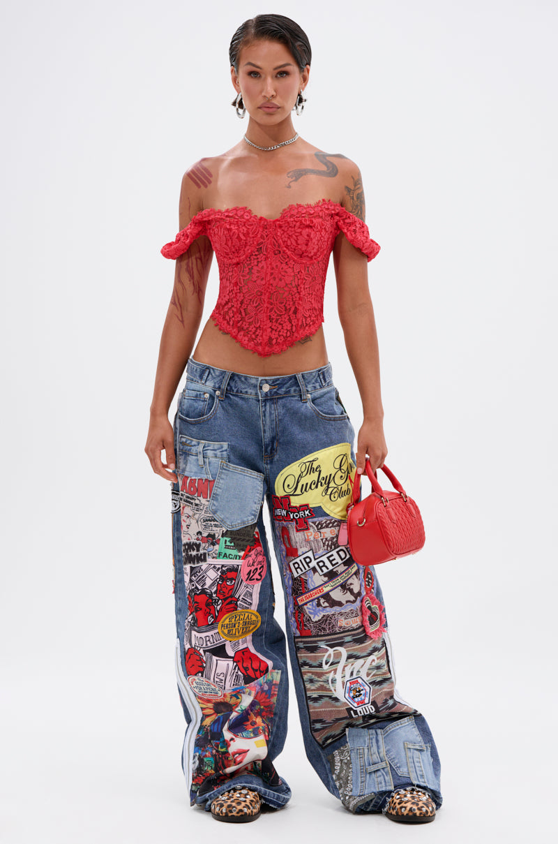 A person stands confidently in the GIRL ON FIRE LACE CORSET, patchwork baggy jeans with colorful graphics, leopard print shoes, and holds a small red handbag against a plain white background.