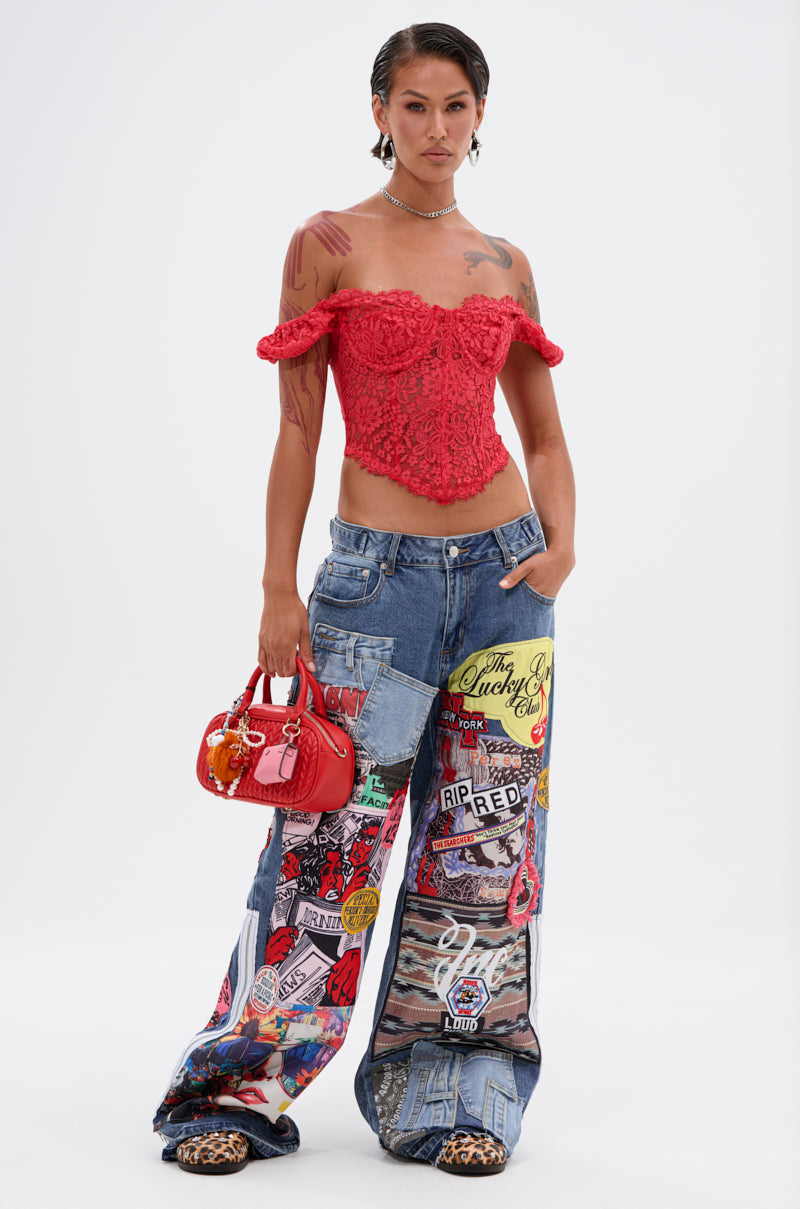 A woman stands against a white background wearing the GIRL ON FIRE LACE CORSET, wide-leg patchwork jeans with colorful graphics and patches, leopard print shoes, and holding a red handbag.