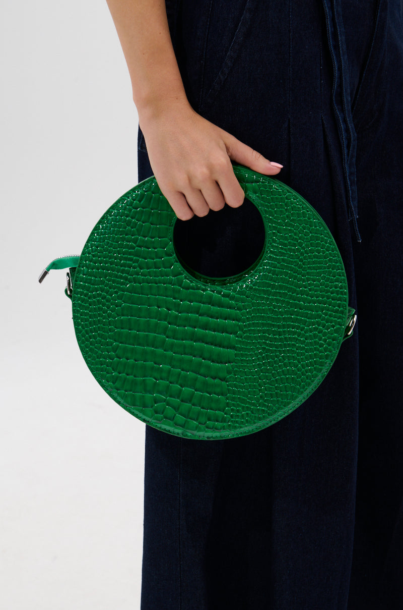 A person dressed in dark blue holds THE CIRCLE PURSE, a textured emerald handbag with a round handle, making it stand out against the simple, light background. 0