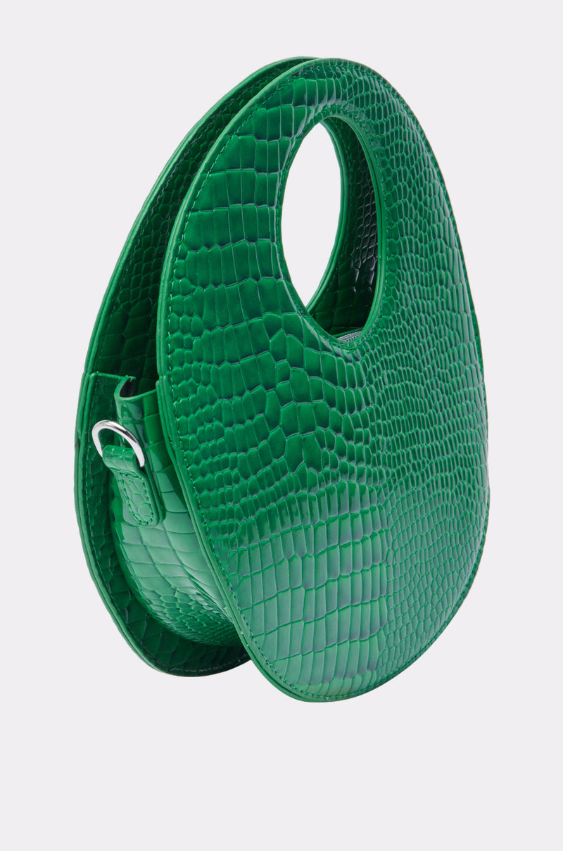 THE CIRCLE PURSE is a green, oval-shaped handbag featuring a crocodile-textured pattern, a circular top handle opening, sleek modern design, and a small side buckle detail.