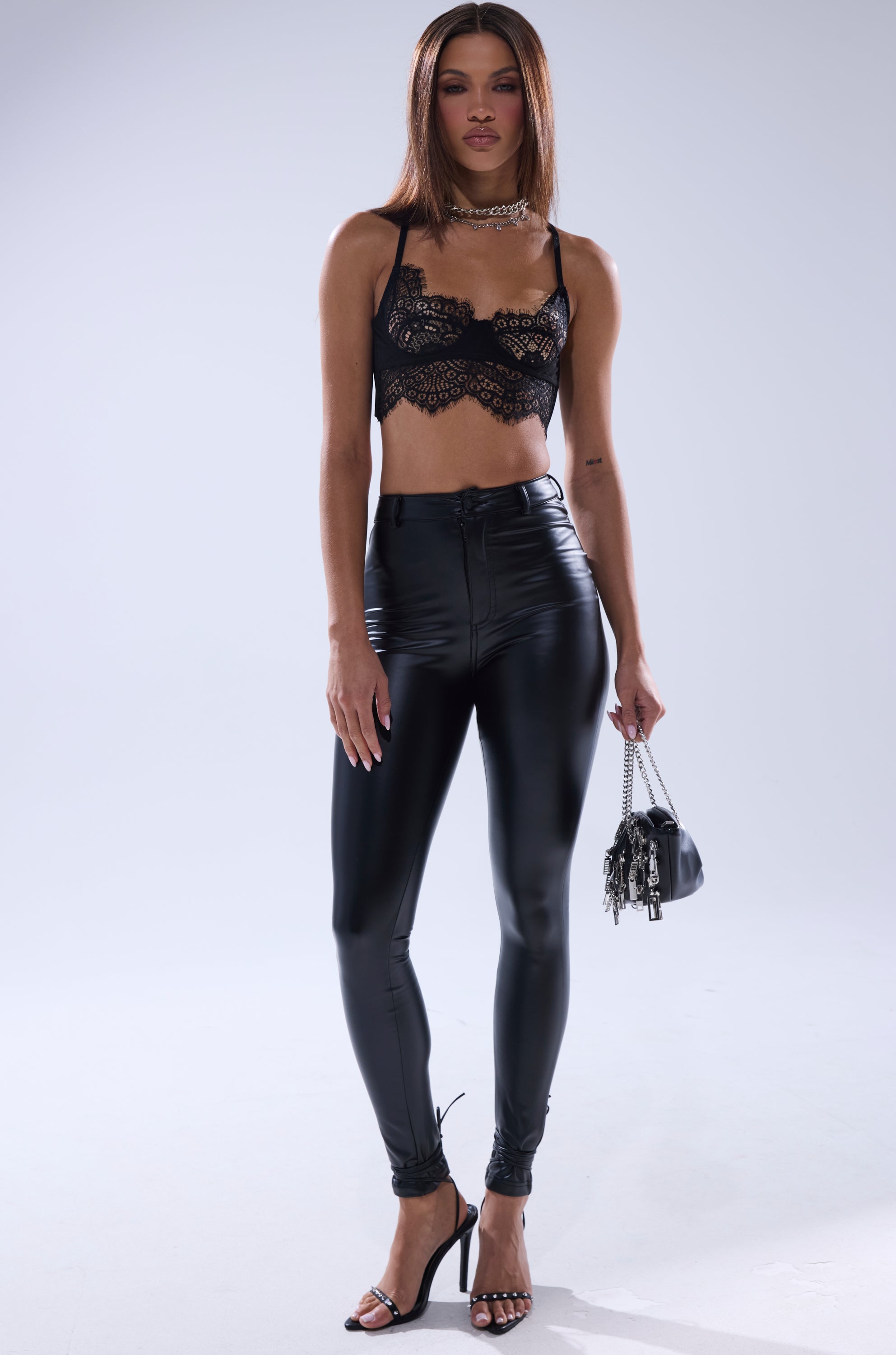 A woman with straight brown hair wears a black lace crop top, TE QUIERO HIGH RISE FAUX LEATHER SKINNY PANTS WITH 4 WAY STRETCH, black strappy heels, a necklace, and holds a small black handbag against a plain background.