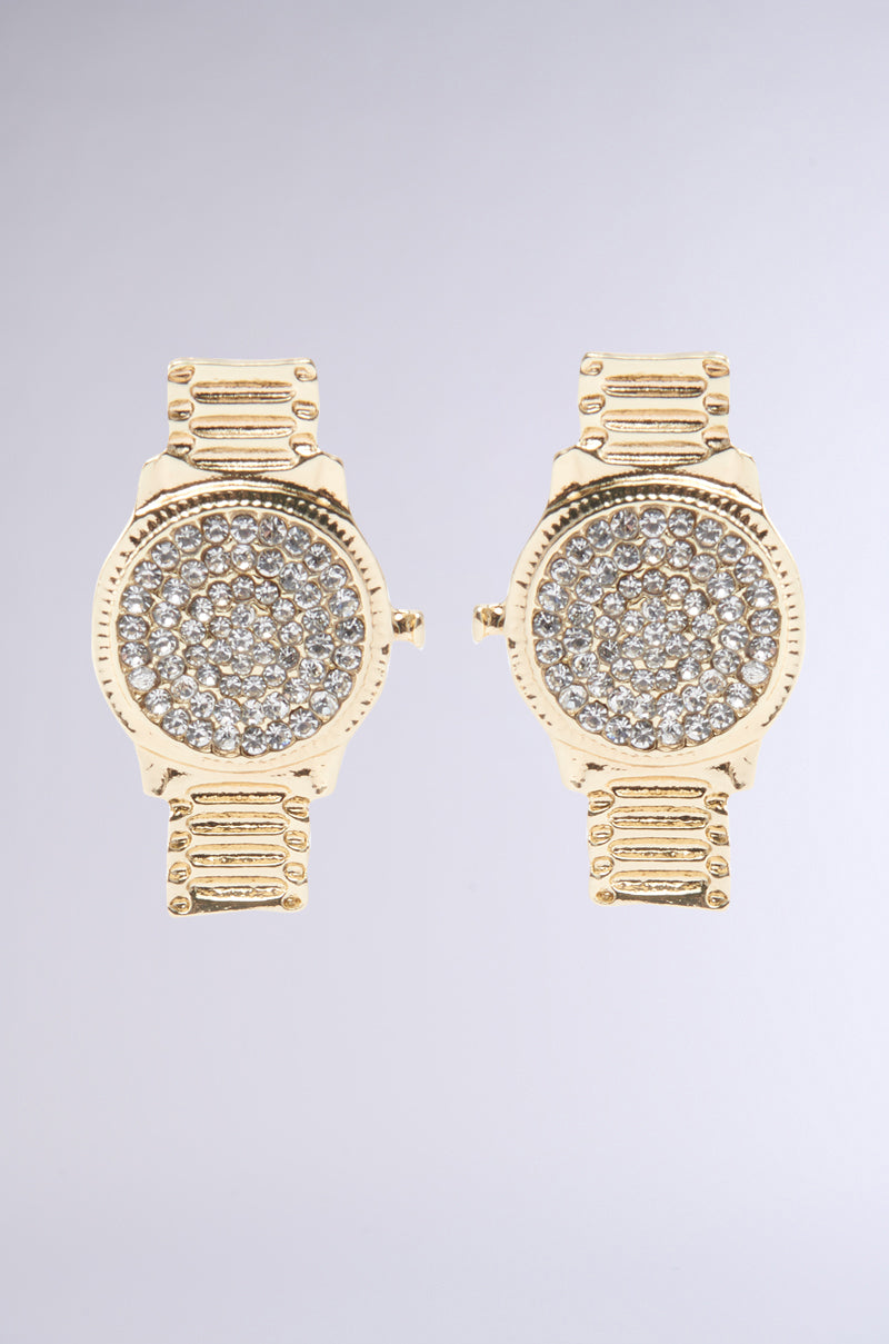 The TAKE YA TIME EARRING features two identical gold, circular watch-inspired pendants encrusted with rhinestones. They boast a textured band-like design and gleam against a neutral gray backdrop.