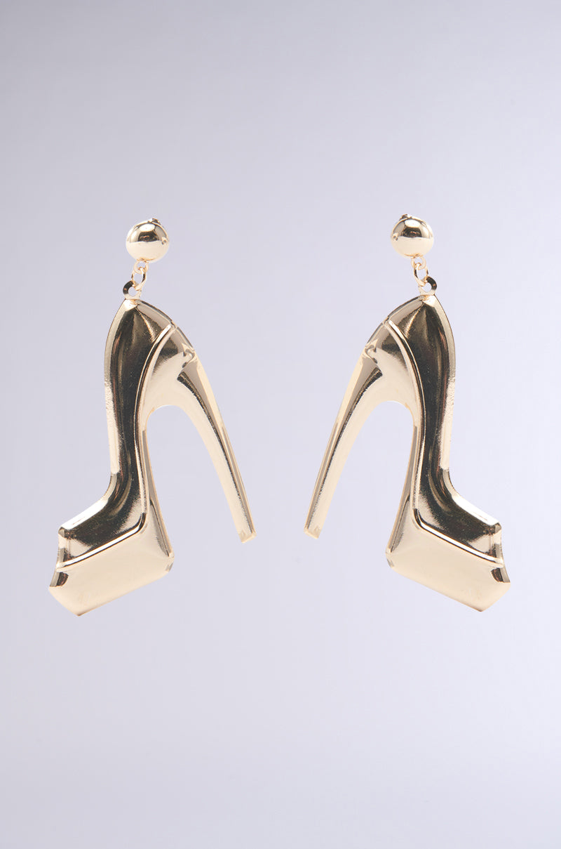 The STOMPERS EARRING, a bold pair of faux gold statement earrings shaped like high-heeled platforms, hang elegantly against a plain light gray background.