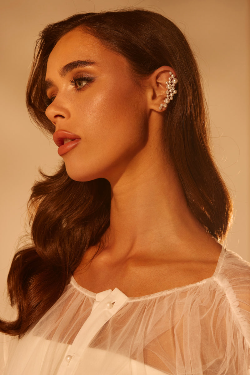A woman with long brown hair poses in warm lighting, wearing a sheer white top and the TOO PREP EARRING—an ornate ear cuff accessory adorned with faux silver pearls—while gazing calmly to the side.