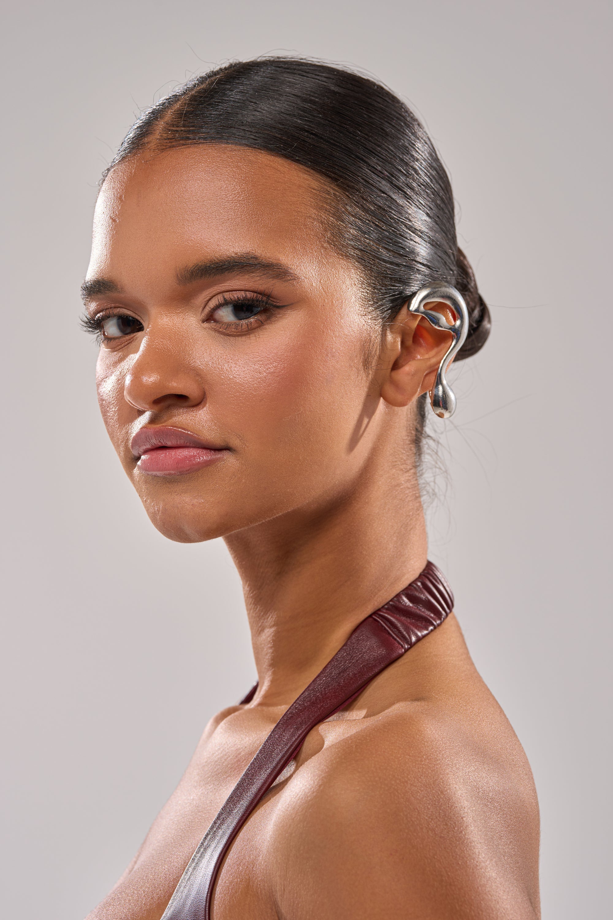 A young woman with smooth, glowing skin and slicked-back dark hair looks over her shoulder, wearing a maroon halter top and the STEF EAR CUFF—a striking silver geometric accessory—against a light grey background. 0