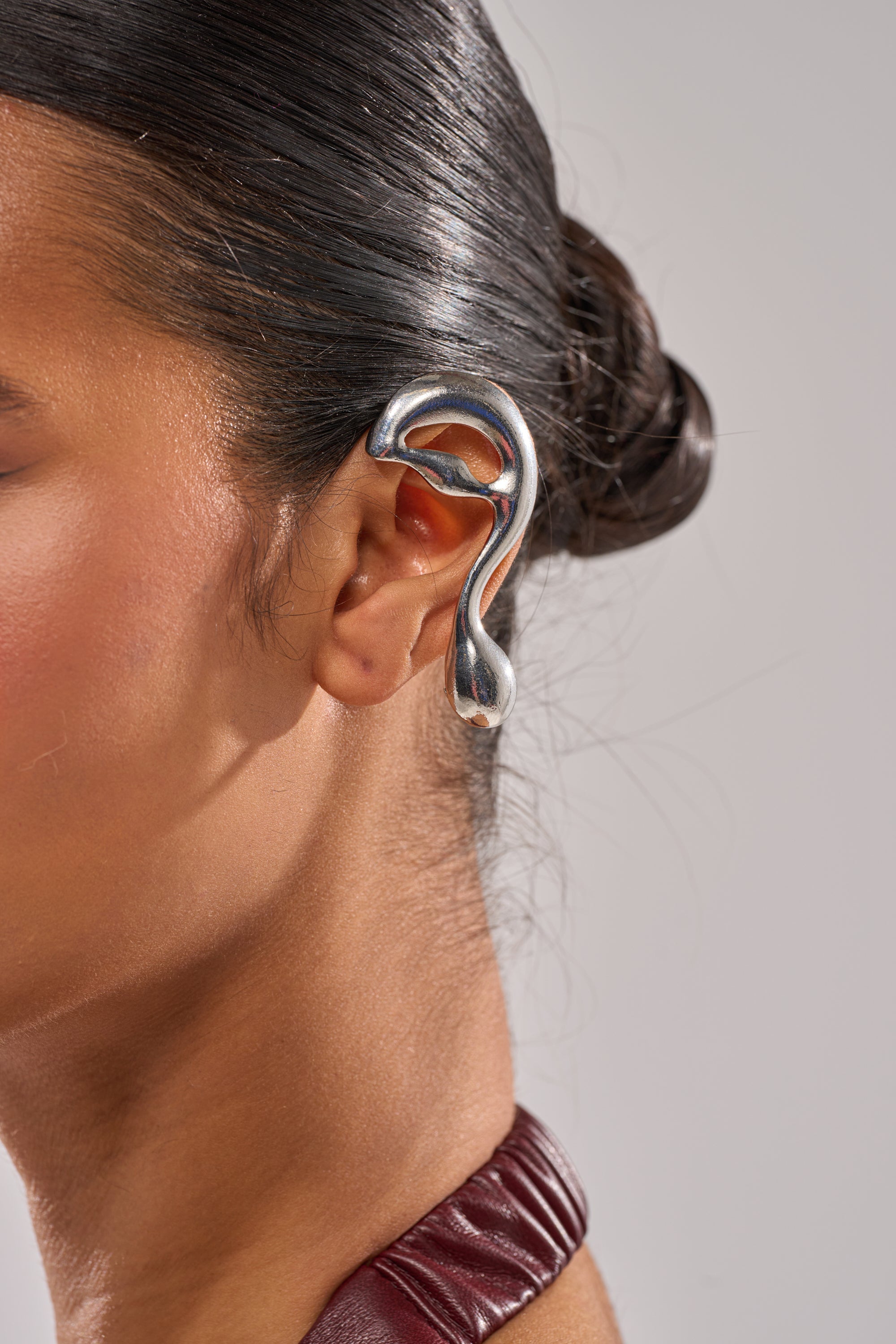 Close-up of a person with dark hair in a sleek bun, showcasing the STEF EAR CUFF—a large, sculptural silver ear cuff with an abstract ear shape. Only the side of the face, ear, and part of the neck are visible. 1