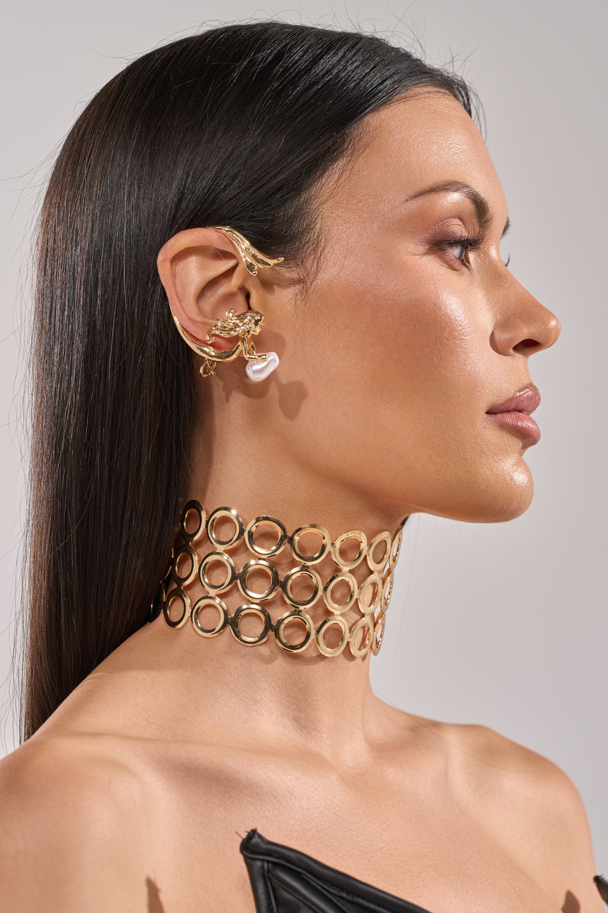 A woman with straight dark hair poses in profile against a neutral background, showcasing her elegant SIREN EAR CUFF—a gold ear cuff paired with a pearl earring and a wide gold choker of interlinked circles. 5