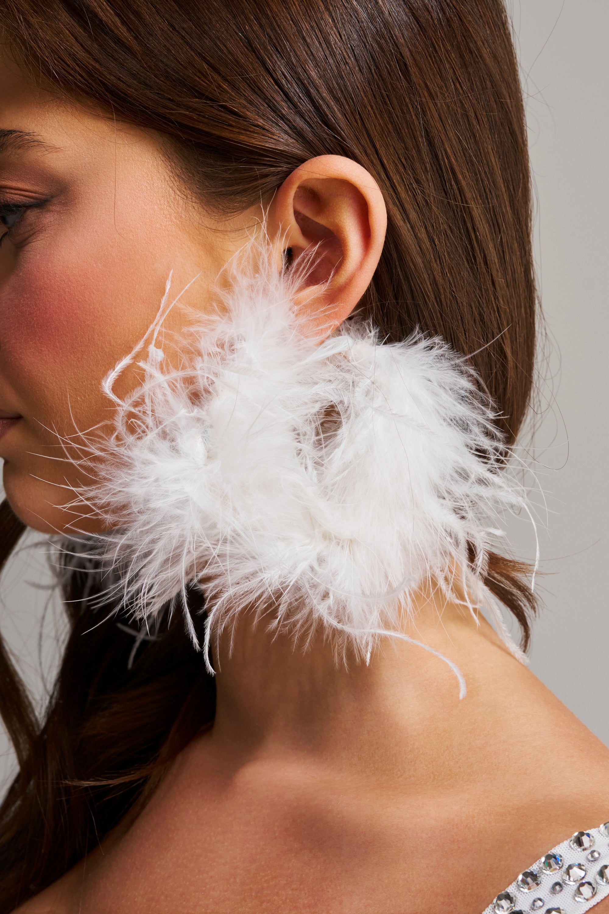 Close-up of a woman wearing the WITH IT FEATHER HOOP EARRING, featuring fluffy white feathers. Her long brown hair is tucked behind her ear, and she sports natural makeup with rosy cheeks against a neutral gray background.