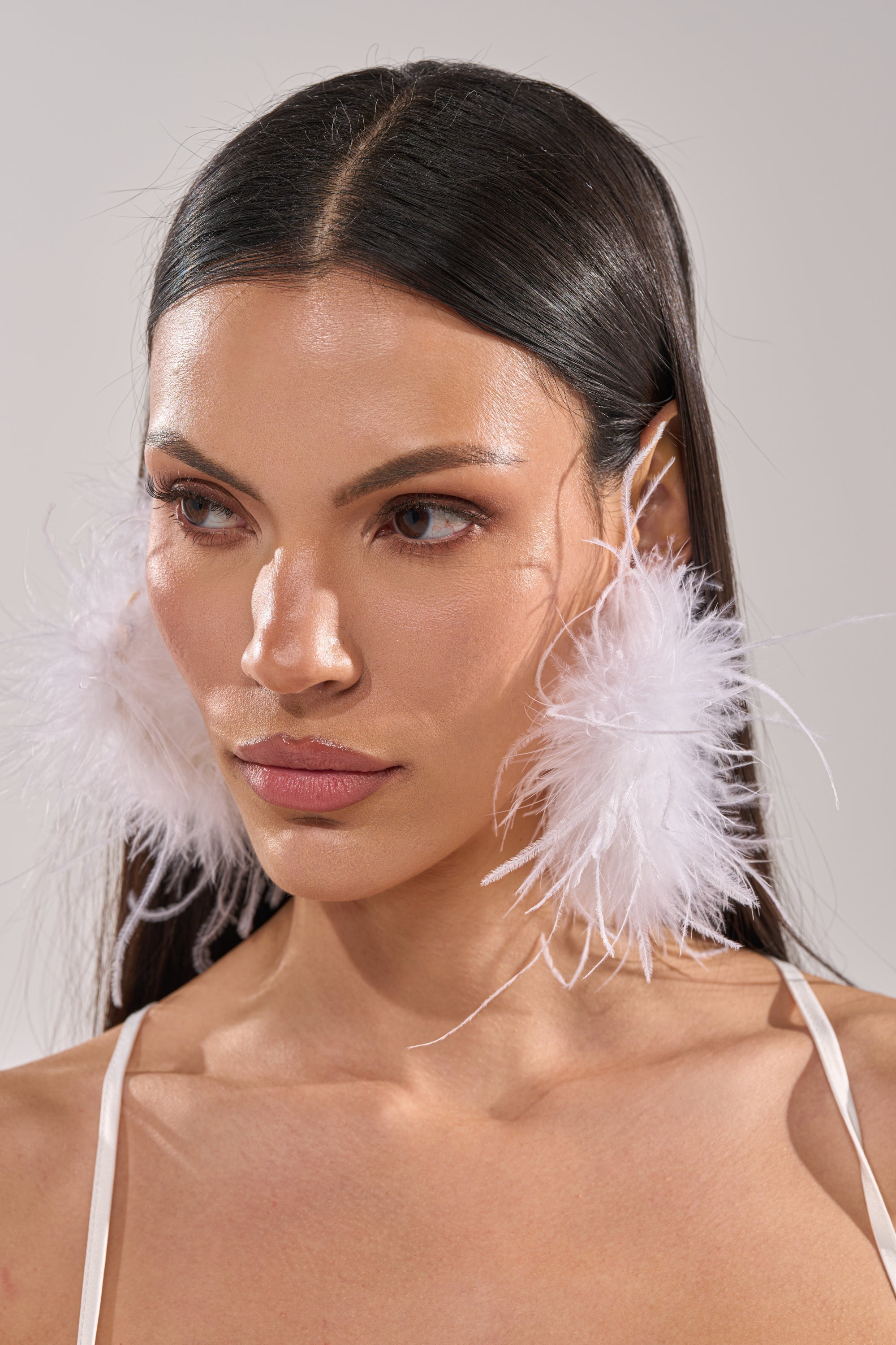 A woman with straight dark hair models the WITH IT FEATHER HOOP EARRING, pairing the bold accessory with a white spaghetti strap top against a neutral background. 0
