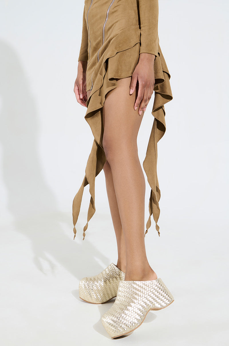 The person is wearing a beige, asymmetrical dress with ruffled edges paired with the AZALEA WANG TEATREE GOLD WOVEN CLOGs, which have a shiny metallic vegan leather finish. The scene is set against a light, neutral background.