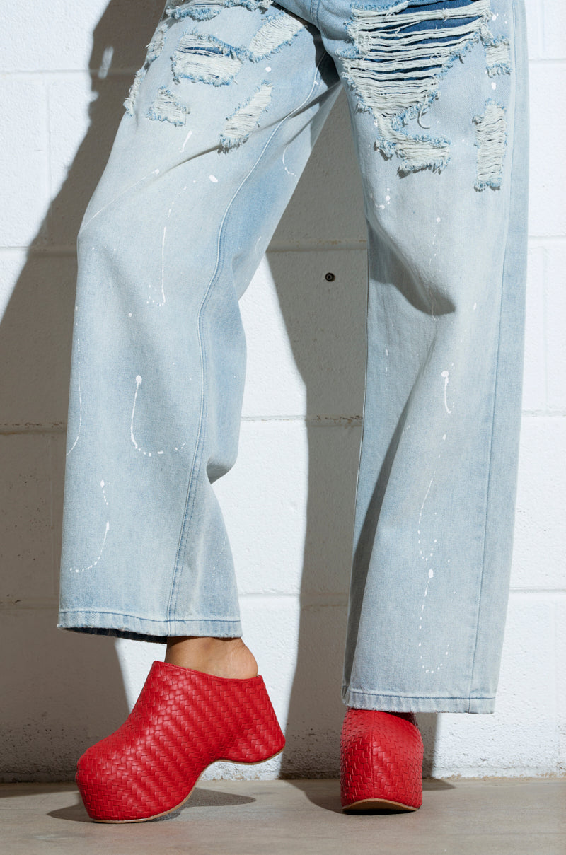 A person is wearing distressed light blue jeans with paint splatters and AZALEA WANG TEATREE RED CLOGS featuring a chunky heel. The photo highlights the lower half of the body against a crisp white brick wall background.