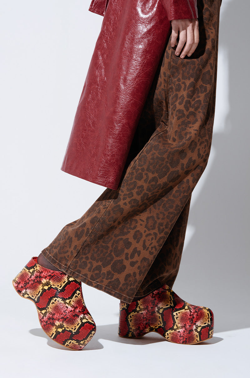 A person dressed in brown leopard-print pants and a red leather coat is standing on tiptoe. They are wearing the AZALEA WANG TEATREE RED SNAKE FLATFORM CLOGS, which feature a colorful snake-skin pattern and a flatform sole, offering both style and support. The background is plain white.