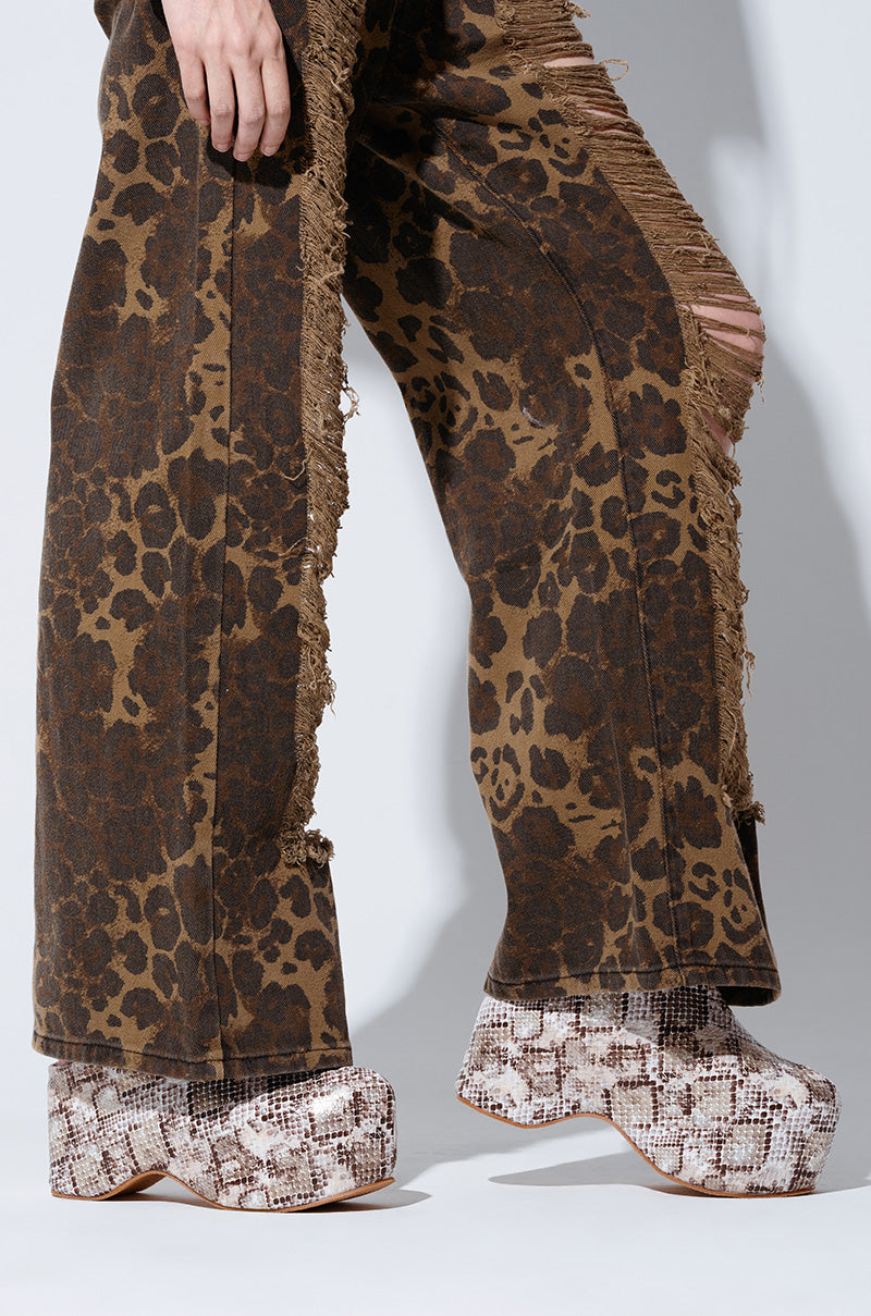 A person stands against a neutral background, showcasing the lower half of their body in wide-leg, leopard-print pants with distressed edges and a pair of AZALEA WANG TEATREE SNAKE FLATFORMS.