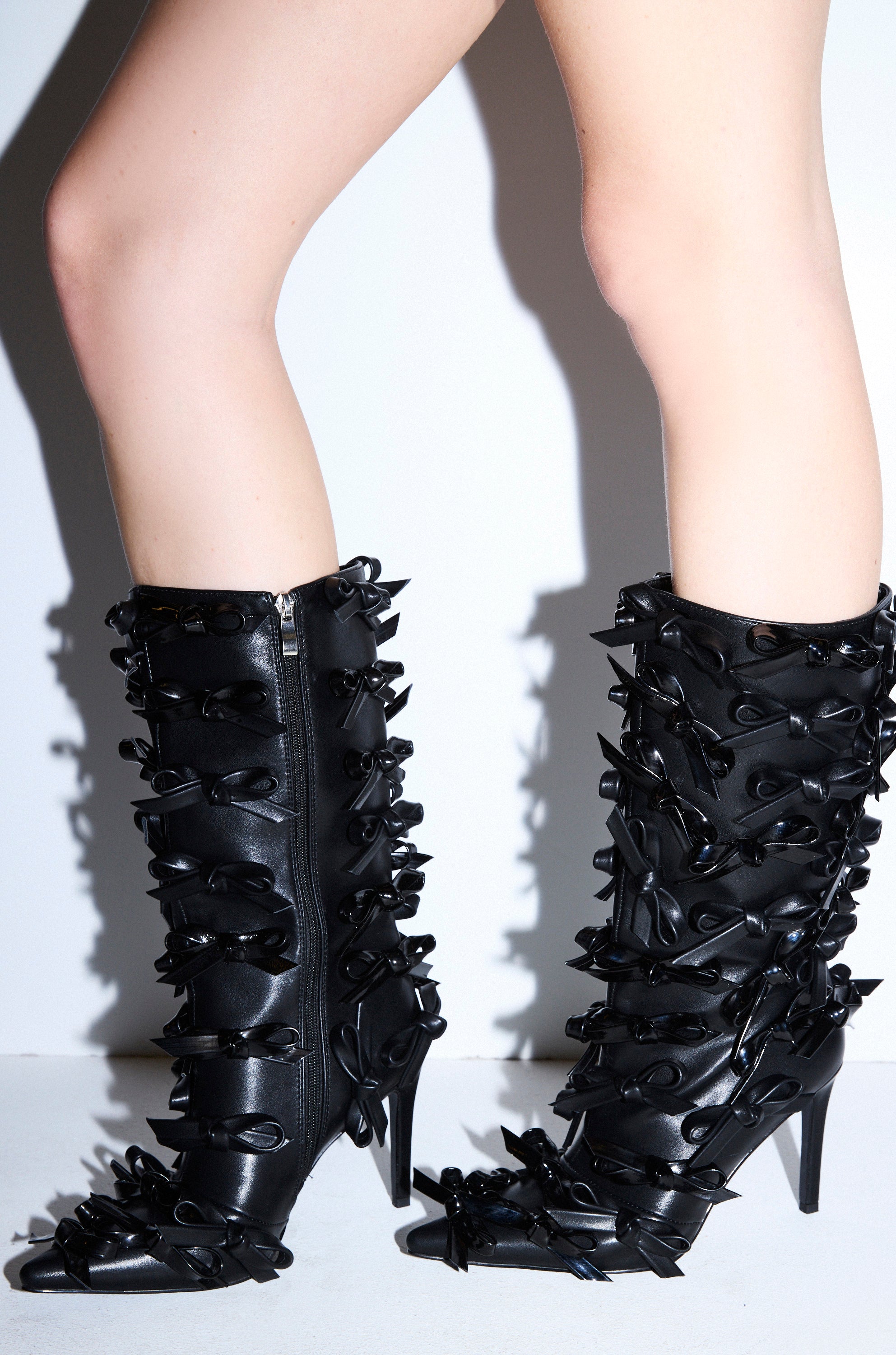 A person wears the AZALEA WANG TELMO BLACK BOW EMBELLISHED BOOTIE—black, knee-high stiletto heel booties with multiple black bows and a convenient side zipper—while standing against a white background.