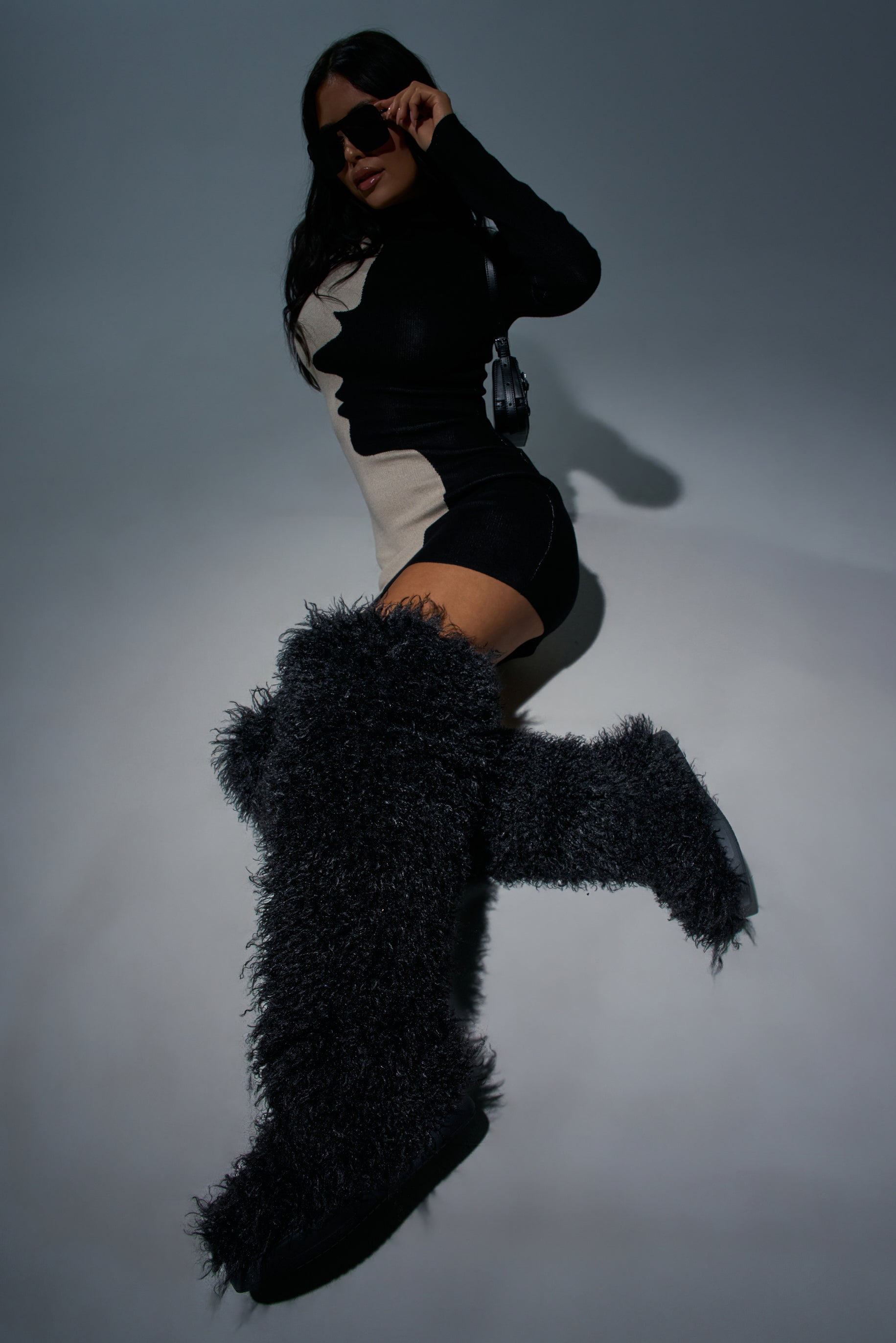 Wearing the AZALEA WANG TEMPLETON BLACK POODLE FAUX FUR OVER THE KNEE BOOT, a person in a black and beige mini dress and sunglasses poses dramatically against a gray background, making a bold statement with their festival footwear.