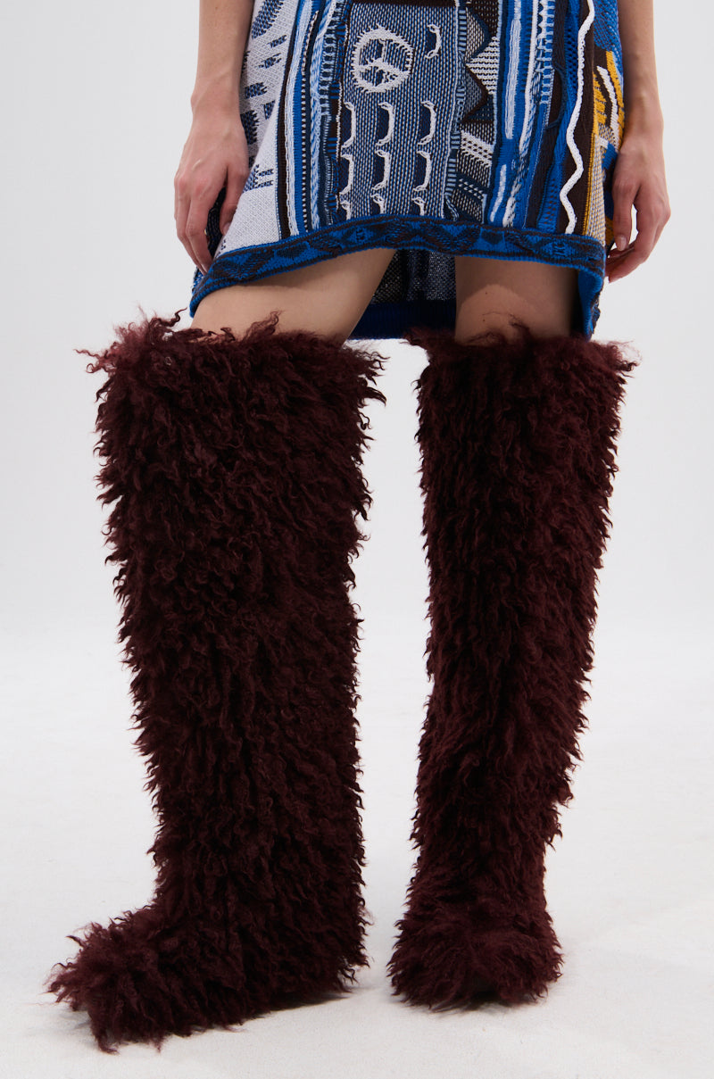 A person stands on a plain white background, wearing a patterned blue skirt with legs visible, styled with AZALEA WANG TEMPLETON BURGUNDY POODLE FUR OVER THE KNEE BOOTS featuring tall, shaggy faux fur that extends above the knees.