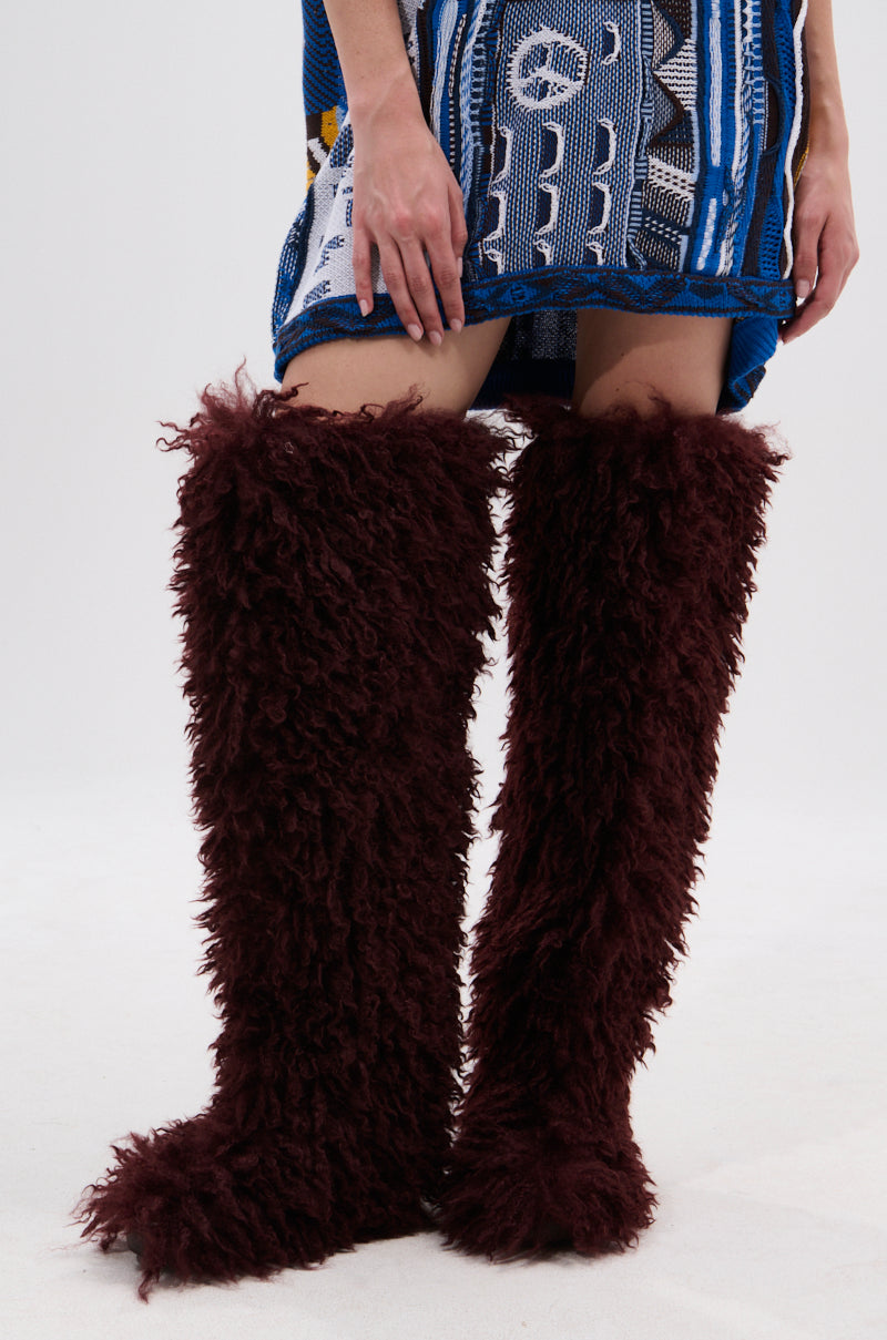 A person wears a short, patterned blue and white dress and AZALEA WANG TEMPLETON BURGUNDY POODLE FUR OVER THE KNEE BOOTS, standing against a plain white background.