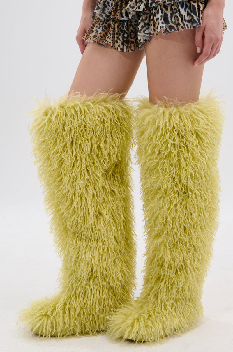 A person wears the AZALEA WANG TEMPLETON LIME POODLE FUR OVER THE KNEE BOOT with a short, ruffled leopard print skirt, standing against a plain white background.