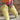A person wearing a colorful sequined dress and AZALEA WANG TEMPLETON Lime Poodle Fur Over The Knee Boots walks on gravel in front of a brick wall, carrying a small pink sparkly purse.