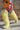 A person wearing a colorful sequined dress and AZALEA WANG TEMPLETON Lime Poodle Fur Over The Knee Boots walks on gravel in front of a brick wall, carrying a small pink sparkly purse.