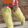 A person wearing a colorful sequined dress and AZALEA WANG TEMPLETON Lime Poodle Fur Over The Knee Boots walks on gravel in front of a brick wall, carrying a small pink sparkly purse.