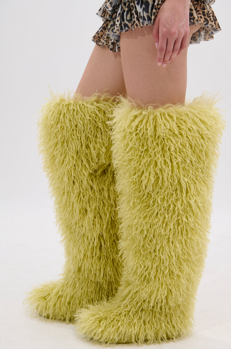 A person wears tall, fuzzy, light yellow AZALEA WANG TEMPLETON LIME POODLE FUR OVER THE KNEE BOOTS with a short leopard print skirt against a plain white background.
