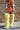 A person stands outdoors against a brick wall, wearing a colorful long-sleeve top and AZALEA WANG TEMPLETON LIME POODLE FUR OVER THE KNEE BOOTS. Their face is not visible in the image.