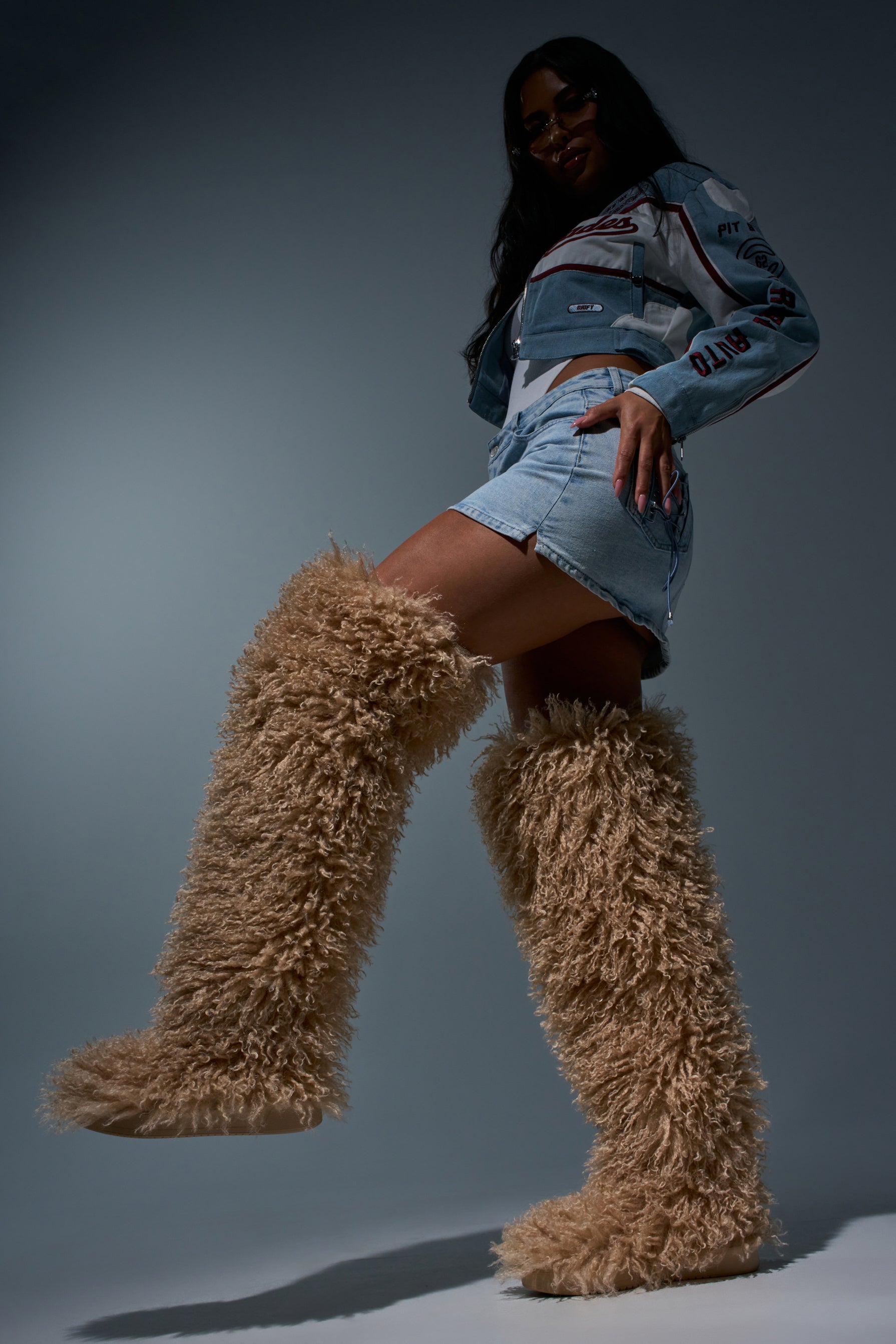 Confidently posing against a dark background, a person styled in sunglasses, a denim jacket, and shorts lifts one leg forward to showcase the cozy-chic AZALEA WANG TEMPLETON NUDE POODLE FUR OVER THE KNEE BOOT—tall tan boots adorned with plush faux poodle fur.