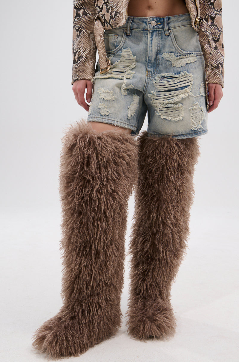 A person wears distressed denim shorts, a snakeskin-patterned top, and AZALEA WANG TEMPLETON TAUPE POODLE FUR OVER THE KNEE BOOTS, standing against a plain white background.