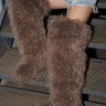 A person sits on metal stairs wearing distressed denim shorts and AZALEA WANG TEMPLETON TAUPE POODLE FUR OVER THE KNEE BOOTS, which cover their legs up to the thighs.