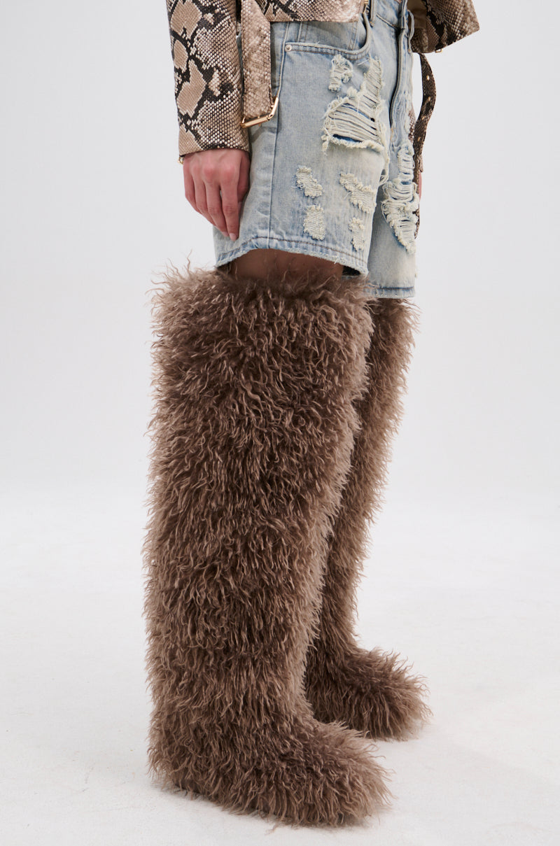 A person in ripped denim shorts, a snakeskin jacket sleeve, and knee-length AZALEA WANG TEMPLETON TAUPE POODLE FUR OVER THE KNEE BOOTS stands against a plain white background.