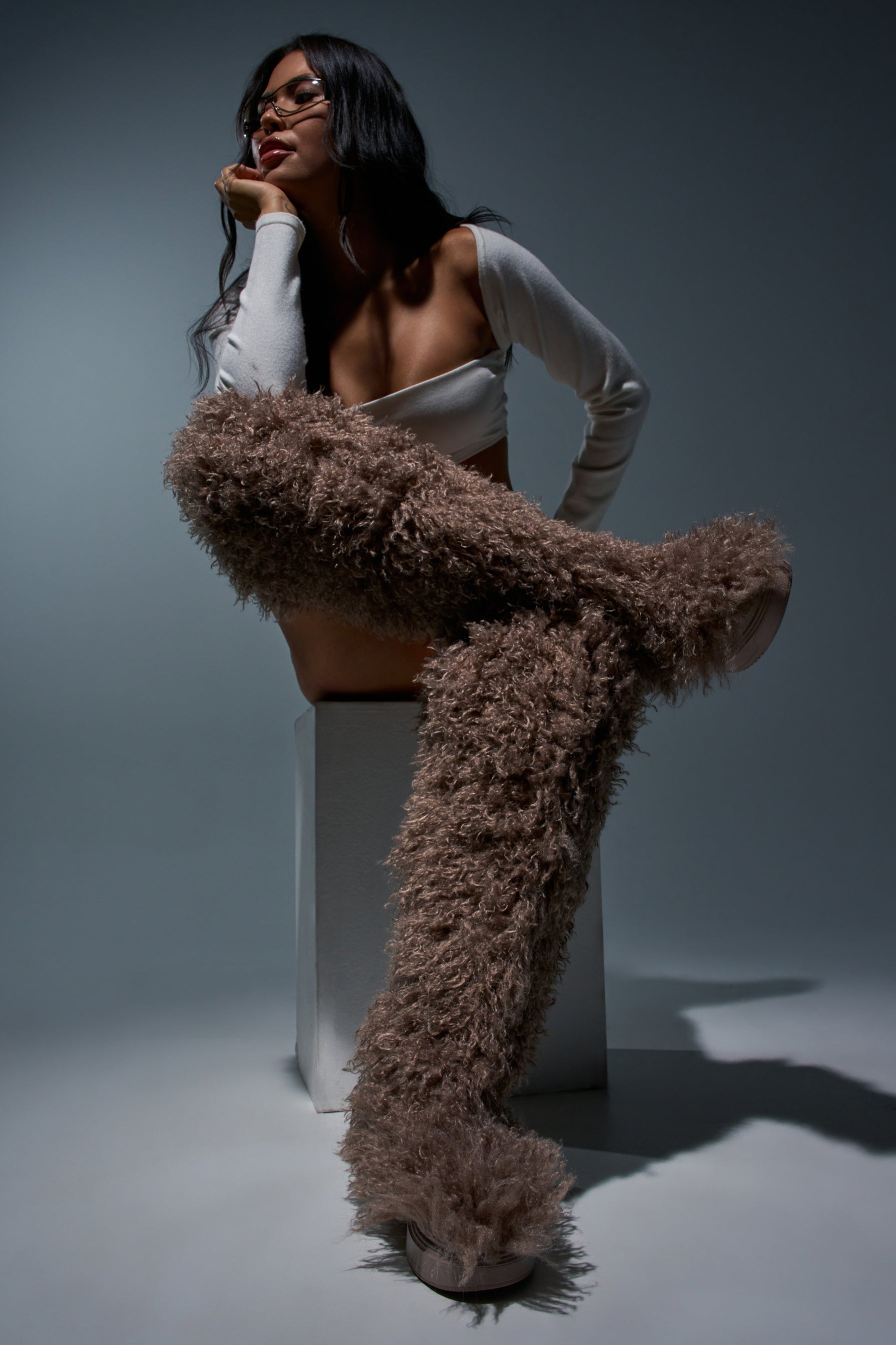 A woman poses seated on a block, wearing a white crop top, long sleeves, glasses, and the AZALEA WANG TEMPLETON TAUPE FAUX POODLE FUR OVER THE KNEE BOOT—striking over-the-knee boots crafted from faux poodle fur that cover her legs almost to the waist. The dramatic lighting with deep shadows perfectly highlights her bold cold-weather styling.