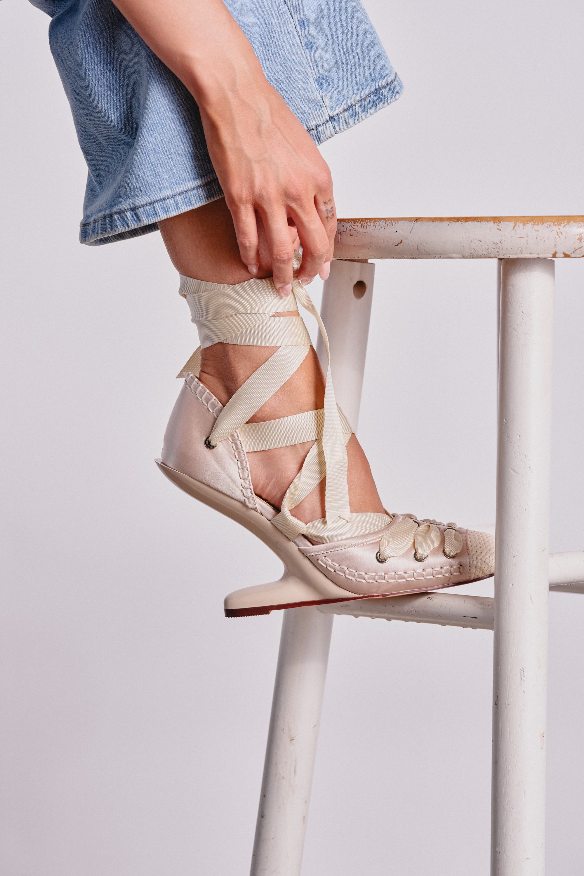 A person in light blue jeans sits on a white wooden stool, tying the ivory lace-up ankle straps of the AKIRA GOLD TENDU BALLET LACE UP NOVELTY HEEL PUMP, which features a square toe and ballet-inspired style. 1