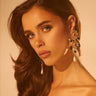 Under warm, golden lighting, a woman with long, wavy brown hair and a serious expression showcases the bold DONATELLA EARRING as she looks into the camera.
