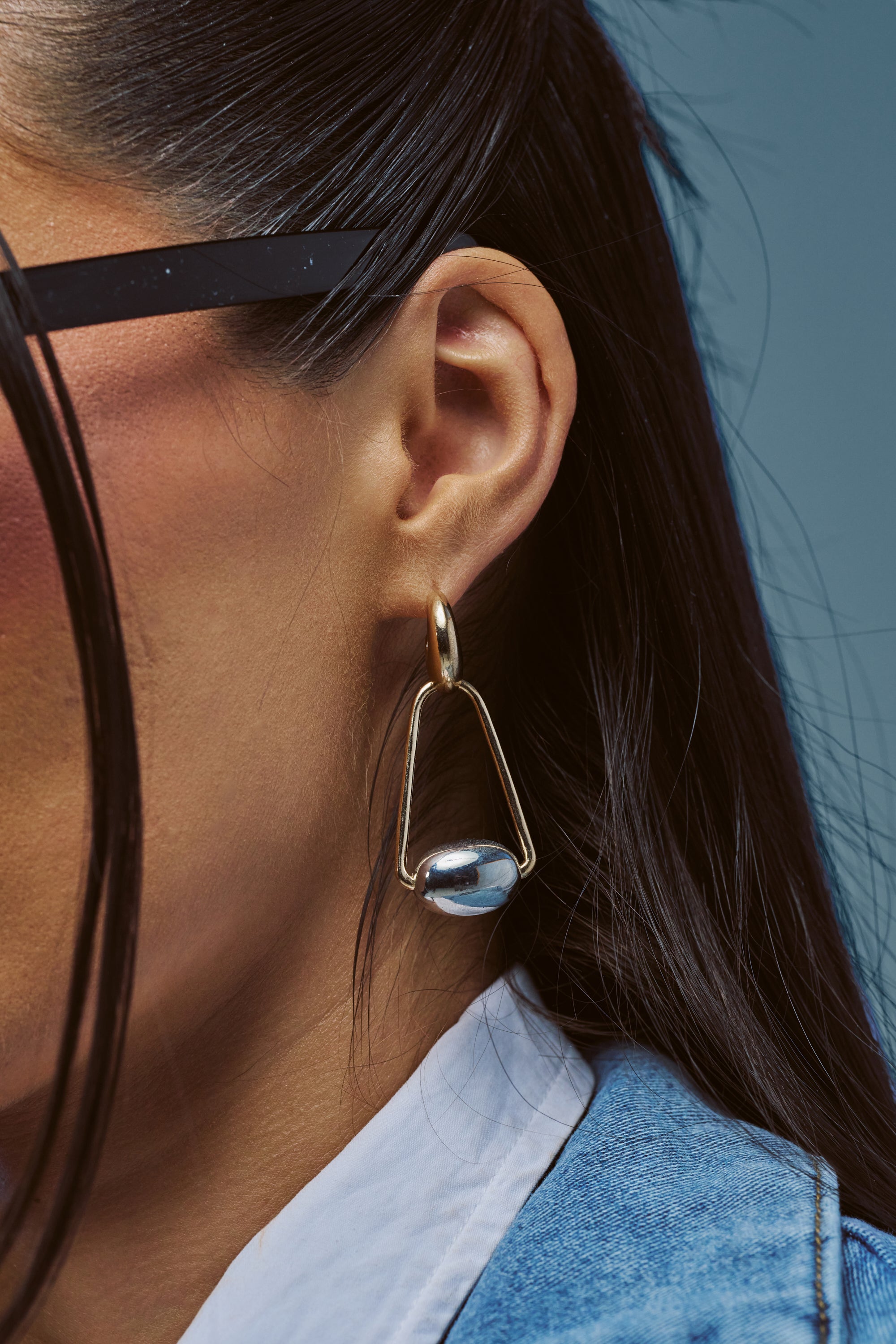 A close-up shows a person with dark hair and glasses, wearing GEOMETRIC DANGLE DROP EARRINGS in gold, paired with a blue denim jacket over a white shirt, set against a blue background.