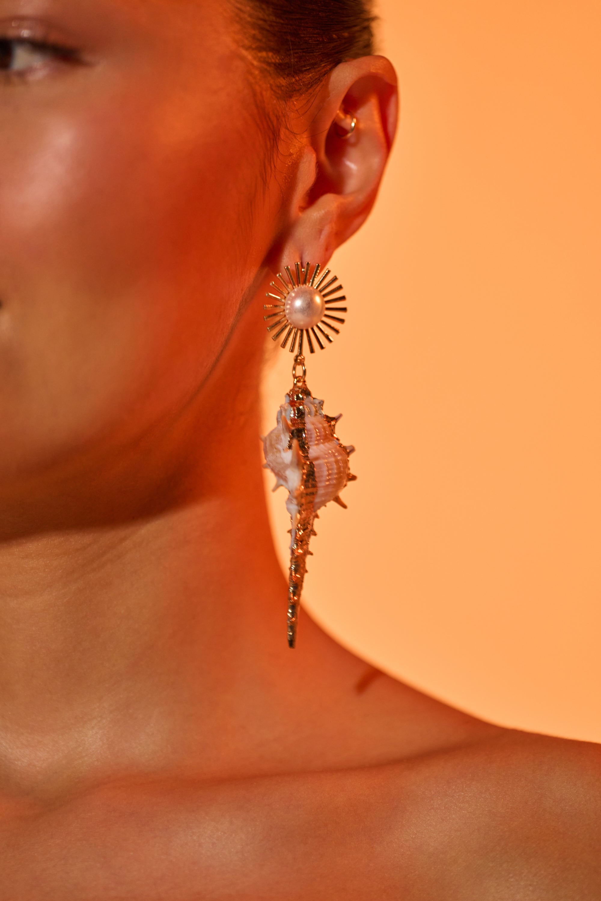 A close-up of a woman’s ear wearing the TO THE SHORE EARRING—striking beach-inspired jewelry with a sunburst design, gold pearl, and hanging seashell—set against a warm orange background. 0