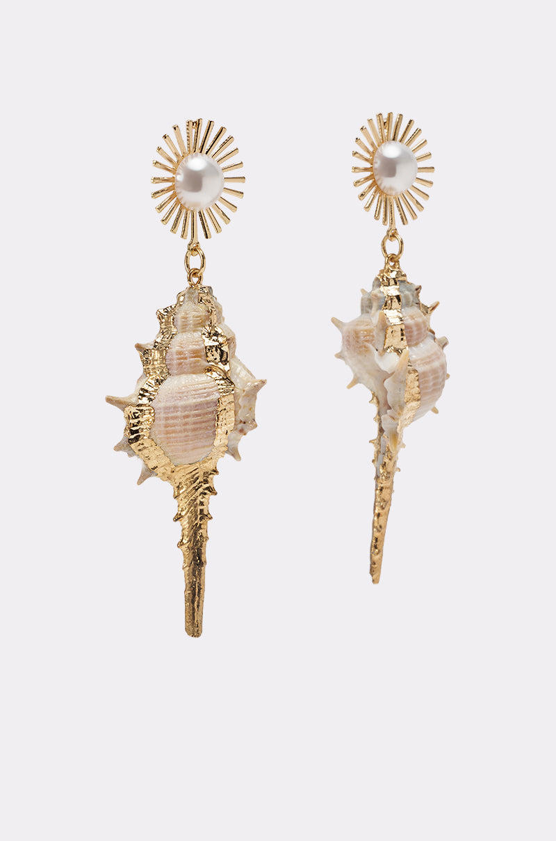The TO THE SHORE EARRING features gold-tone seashells with dangling conch shells and a pearl center at the top, accented by a gold metallic sunburst design. Set against a plain, light-colored background. 2