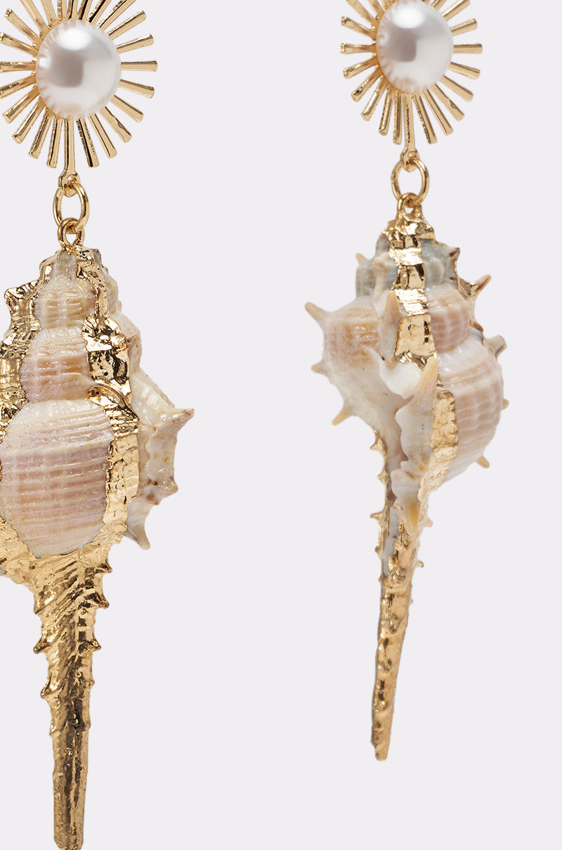 The TO THE SHORE EARRING features seashell charms with gold accents, hanging from sunburst studs topped with pearl centers—beach-inspired earrings that shine with gold-toned elegance against a plain backdrop. 3