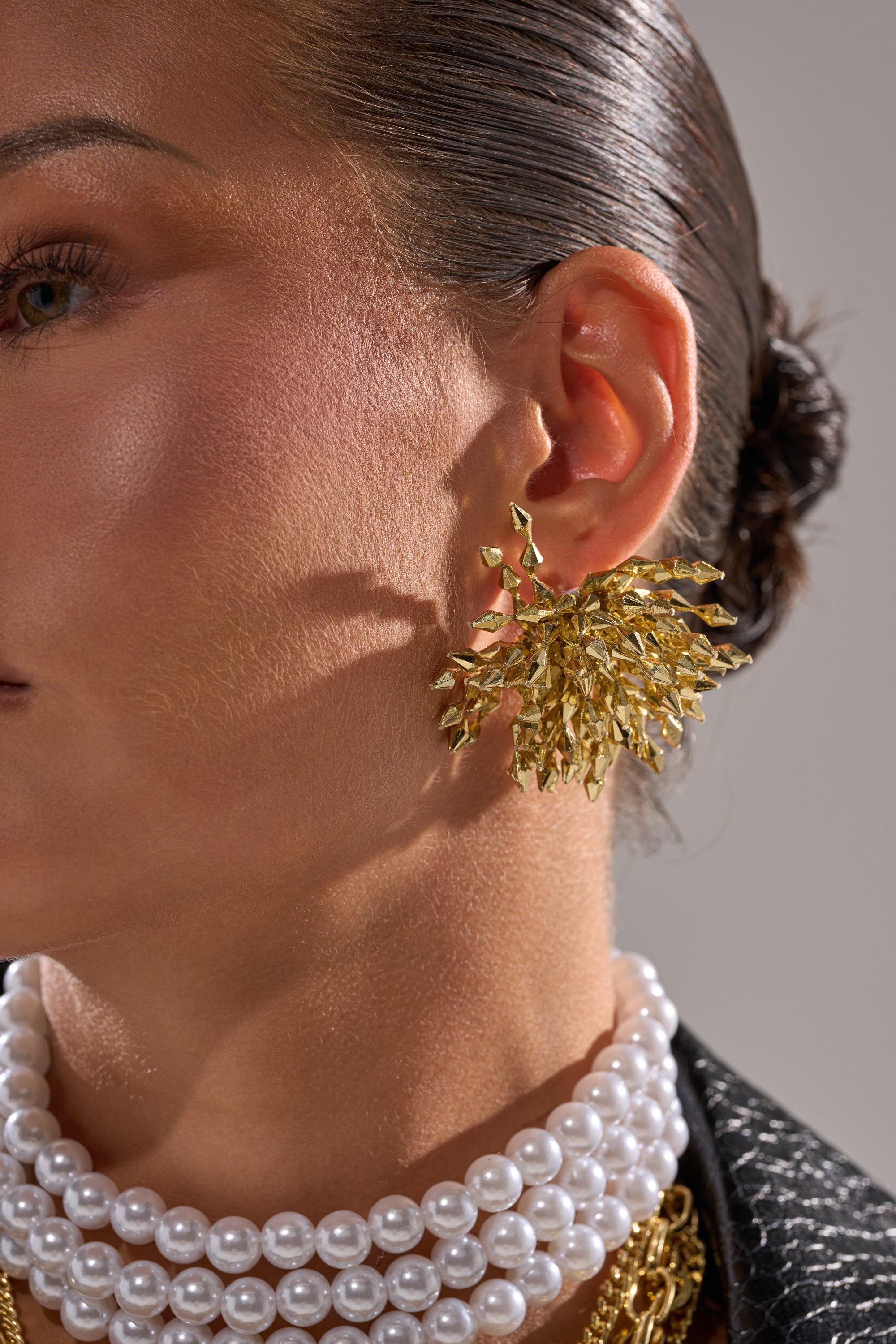 Close-up of a woman's face wearing the EDGY VIBE EARRING, paired with layered pearl necklaces and a black textured outfit. Her hair is neatly slicked back in a bun. 4