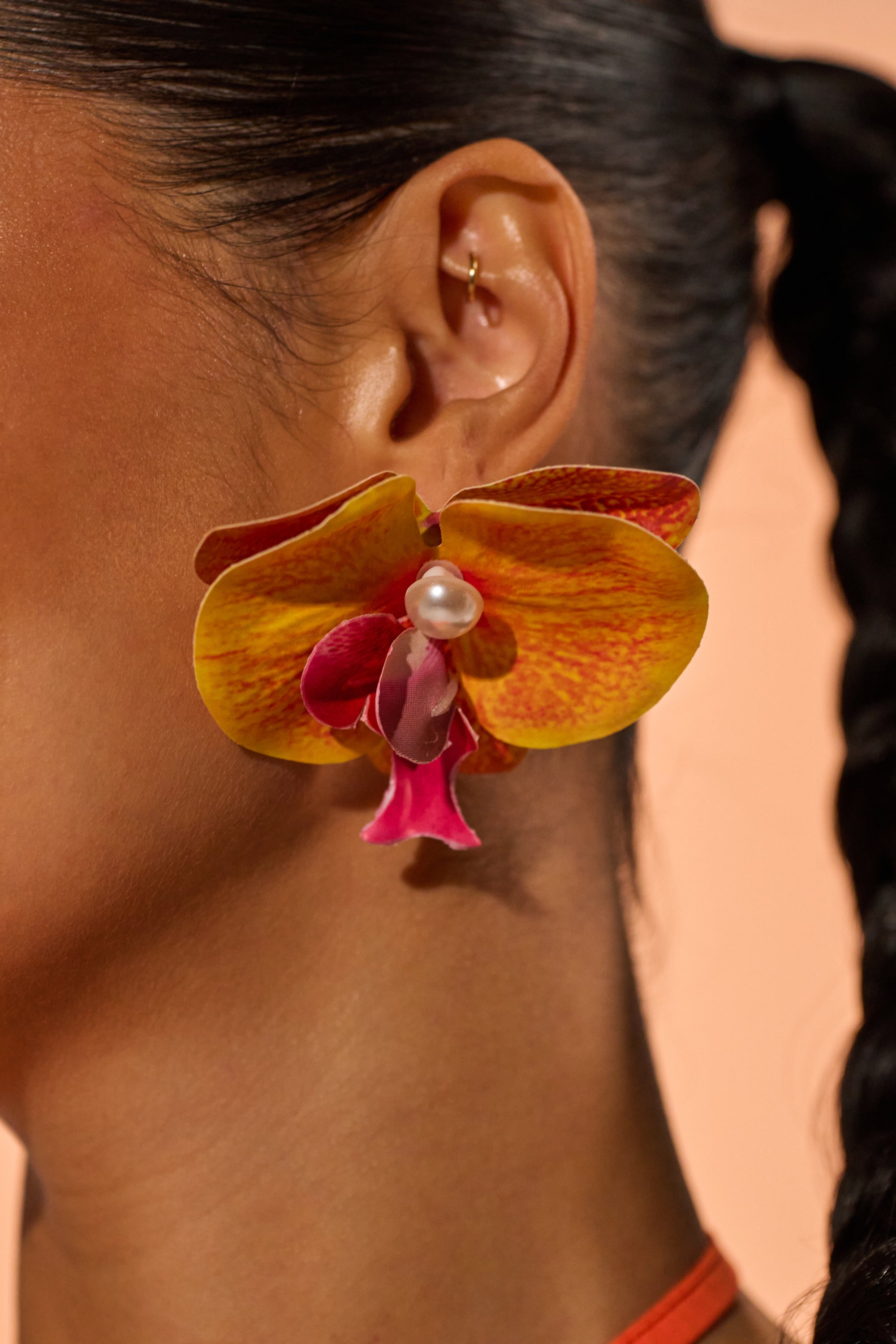 Close-up of a person wearing the NEW SEASON EARRING—an oversized botanical piece shaped like a vibrant yellow and pink tropical flower with a faux pearl center. A small gold hoop decorates the upper ear, and dark hair is styled in a side braid.